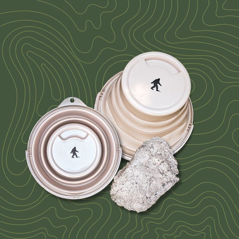 Peut inclure: Gobelets de voyage pliables beiges avec des silhouettes de Bigfoot noires sur les couvercles. Un gobelet est ouvert. Une roche argent&eacute;e textur&eacute;e est &eacute;galement pr&eacute;sente. Fond vert avec une carte topographique.