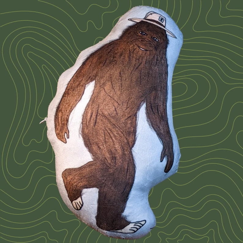 Op de afbeelding: Een pluche speelgoed in de vorm van Bigfoot met een rangerhoed. Het speelgoed is bruin en wit met een pluche textuur.