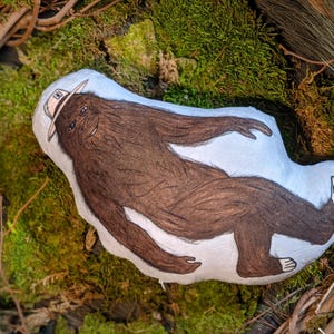 Op de afbeelding: Een bruin pluche speelgoed in de vorm van een Bigfoot met een rangerhoed. Het speelgoed ligt op een bed van groen mos.