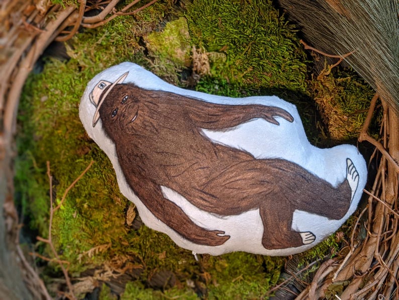 Op de afbeelding: Een bruin pluche speelgoed in de vorm van een Bigfoot met een rangerhoed. Het speelgoed ligt op een bed van groen mos.