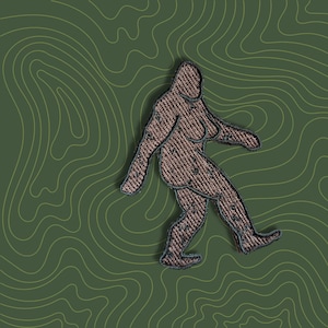 Könnte beinhalten: Ein gestickter Aufnäher, der einen Bigfoot zeigt, der auf einem grünen Hintergrund mit topografischem Muster geht.