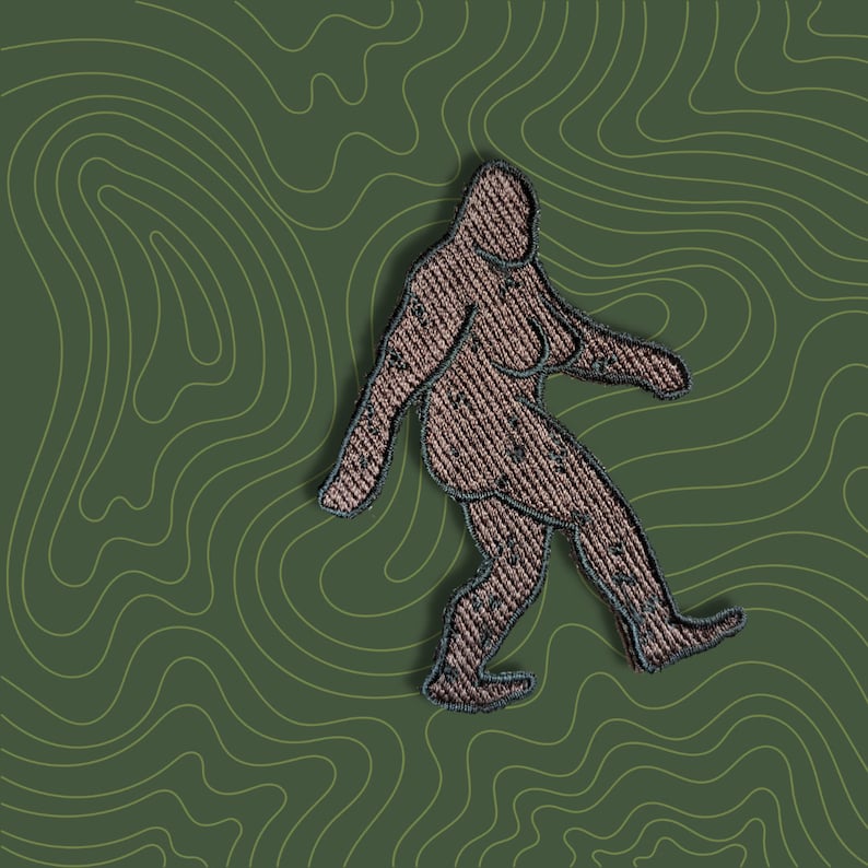 K&ouml;nnte beinhalten: Ein gestickter Aufn&auml;her, der einen Bigfoot zeigt, der auf einem gr&uuml;nen Hintergrund mit topografischem Muster geht.
