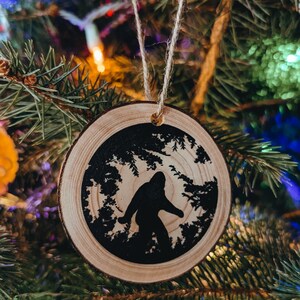K&ouml;nnte beinhalten: Ein runder Weihnachtsbaumschmuck aus Holz mit einer Silhouette von Bigfoot, der durch einen Wald geht. Der Schmuck ist aus rustikalem Holz.