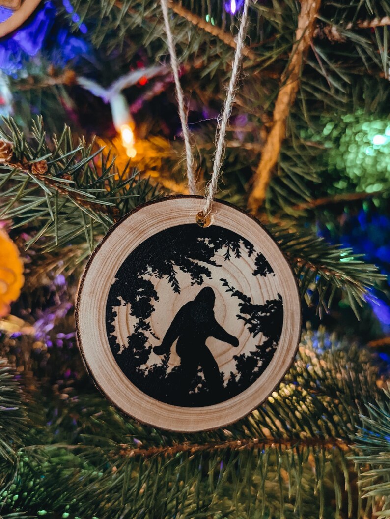 K&ouml;nnte beinhalten: Ein runder Weihnachtsbaumschmuck aus Holz mit einer Silhouette von Bigfoot, der durch einen Wald geht. Der Schmuck ist aus rustikalem Holz.