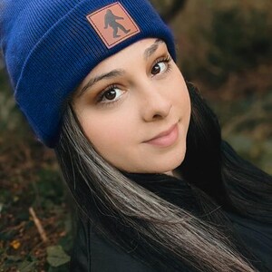 Pu&ograve; includere: Una donna indossa un berretto blu con una toppa in pelle marrone che presenta la silhouette di un Bigfoot. Il berretto &egrave; realizzato in un morbido tessuto a maglia. La donna ha i capelli lunghi e scuri con ciocche grigie.