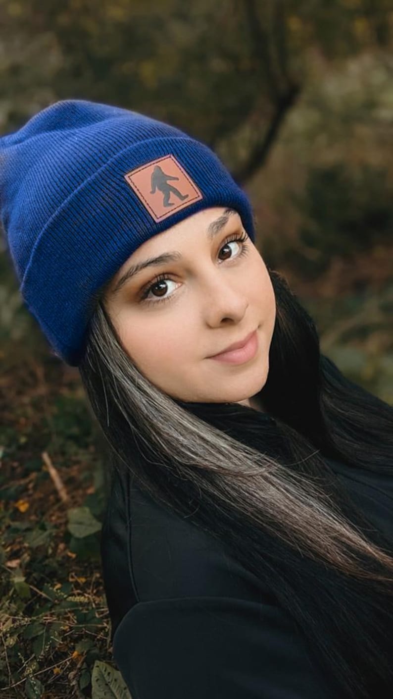 Pu&ograve; includere: Una donna indossa un berretto blu con una toppa in pelle marrone che presenta la silhouette di un Bigfoot. Il berretto &egrave; realizzato in un morbido tessuto a maglia. La donna ha i capelli lunghi e scuri con ciocche grigie.