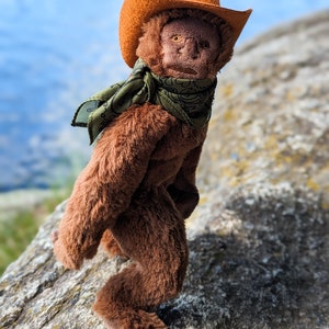 May include: A brown plush monkey wearing an orange cowboy hat with a green and black bandana. The monkey is standing on a rock.