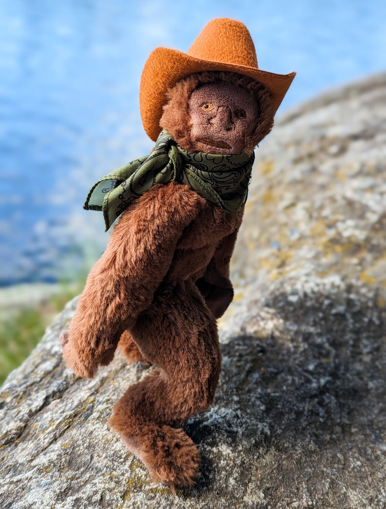 May include: A brown plush monkey wearing an orange cowboy hat with a green and black bandana. The monkey is standing on a rock.