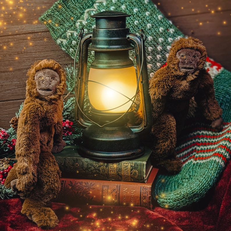 May include: Two brown plush monkey toys sit on a stack of books with a vintage oil lamp in the background. The lamp is lit and casting a warm glow. The monkeys are sitting on a red velvet surface with a green and white knitted blanket in the background.