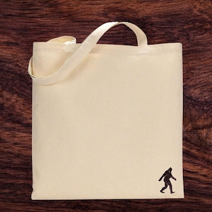 Op de afbeelding: Een natuurlijke canvas draagtas met een bruin silhouet van een Bigfoot die in de rechterbenedenhoek loopt.