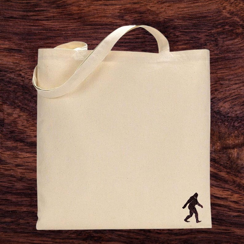 Op de afbeelding: Een natuurlijke canvas draagtas met een bruin silhouet van een Bigfoot die in de rechterbenedenhoek loopt.