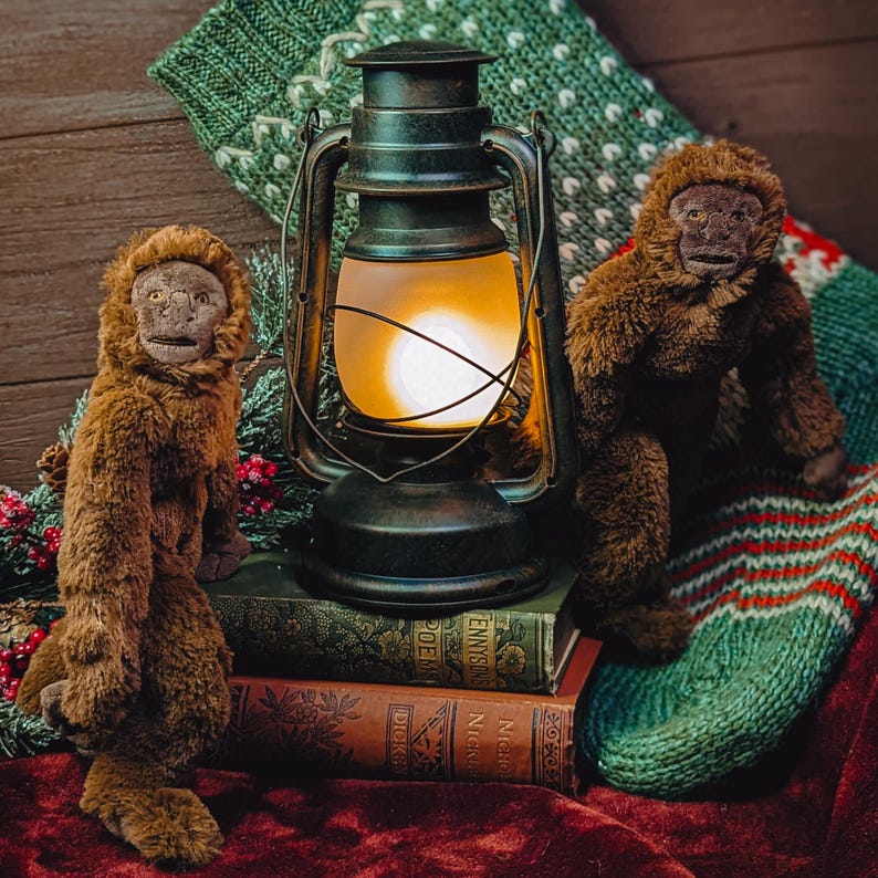 May include: Two brown plush monkey toys sit next to a vintage oil lamp with a glowing light bulb. The lamp is on top of a stack of old books. The monkeys and lamp are in front of a green and white knitted stocking.