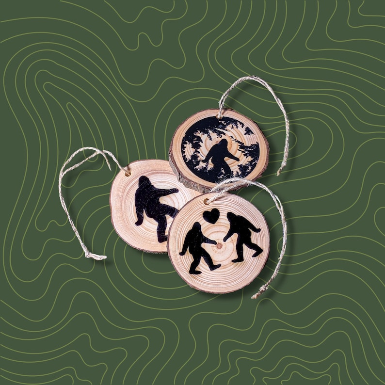 K&ouml;nnte beinhalten: Drei runde Holzornamente mit einer Schnur zum Aufh&auml;ngen. Jedes Ornament zeigt eine Silhouette einer Bigfoot-Kreatur. Ein Ornament zeigt einen einzelnen Bigfoot, ein anderes zeigt zwei Bigfoot, die ein Herz halten, und das dritte zeigt einen Bigfoot, der durch einen Wald geht.