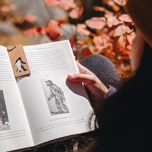 Op de afbeelding: Een houten bladwijzer met een Bigfoot-silhouet ligt in een open boek. Het boek toont tekst en een zwart-wit illustratie van een figuur. De achtergrond is een vage herfstbladeren.