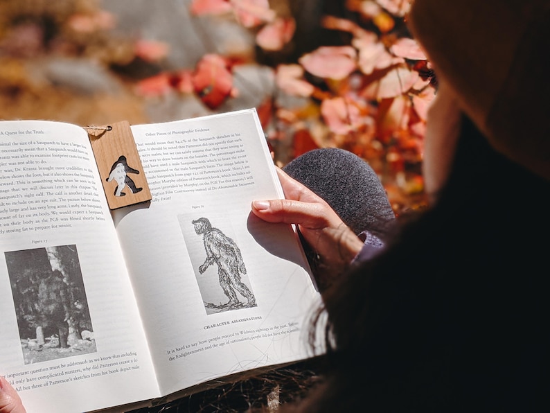 Op de afbeelding: Een houten bladwijzer met een Bigfoot-silhouet ligt in een open boek. Het boek toont tekst en een zwart-wit illustratie van een figuur. De achtergrond is een vage herfstbladeren.