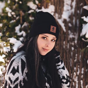 Pu&ograve; includere: Una donna indossa un berretto di lana nero con una toppa in pelle marrone che presenta la sagoma di Bigfoot. Indossa un maglione a motivi bianco e nero e ha lunghi capelli neri.