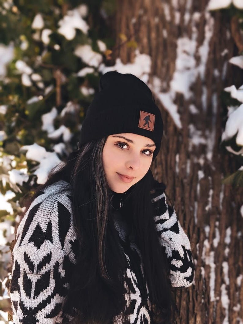 Pu&ograve; includere: Una donna indossa un berretto di lana nero con una toppa in pelle marrone che presenta la sagoma di Bigfoot. Indossa un maglione a motivi bianco e nero e ha lunghi capelli neri.