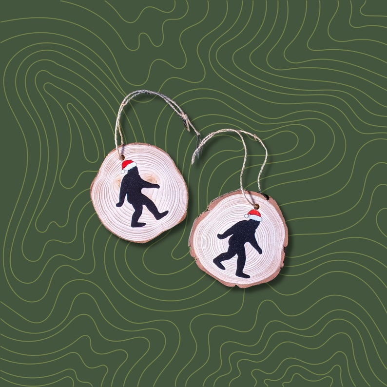 Op de afbeelding: Twee houten ornamenten in de vorm van cirkels met een bruin houtnerfpatroon. Elk ornament heeft een zwart silhouet van een wezen dat een rode kerstmanmuts draagt. De ornamenten hangen aan een touwtje.