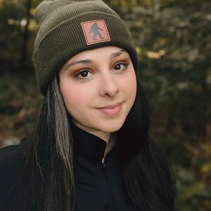 Pu&ograve; includere: Una donna indossa un berretto verde scuro con una toppa in pelle marrone che presenta la silhouette di un Bigfoot. Il berretto &egrave; realizzato in un morbido tessuto a maglia. La donna ha i capelli lunghi e scuri e indossa una camicia nera a collo alto.