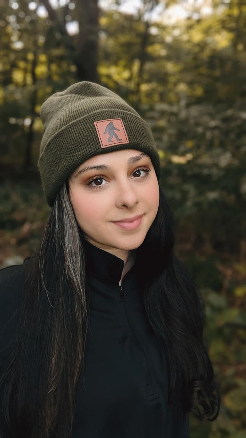 Pu&ograve; includere: Una donna indossa un berretto verde scuro con una toppa in pelle marrone che presenta la silhouette di un Bigfoot. Il berretto &egrave; realizzato in un morbido tessuto a maglia. La donna ha i capelli lunghi e scuri e indossa una camicia nera a collo alto.
