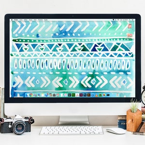 May include: A white desktop computer with a blue and green geometric pattern wallpaper. The computer is on a white desk with a keyboard, mouse, camera, books, and a small potted plant.