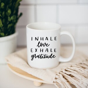 May include: White ceramic coffee mug with black text that reads "INHALE love EXHALE gratitude". The mug is sitting on a wooden coaster with a white fringed cloth.