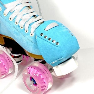 Puede incluir: Primer plano de un patín de ruedas azul claro con cordones blancos y ruedas rosas. El patín tiene una textura similar a la gamuza y ojales plateados. Las ruedas son transparentes con un interior rosa.