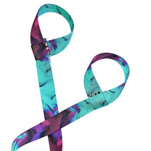 Puede incluir: Una correa de yoga turquesa y morada con dos lazos. La correa está hecha de un material suave y elástico y está diseñada para ayudarte a mejorar tu flexibilidad y rango de movimiento.