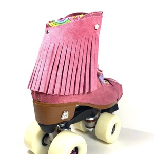 May include: Pink suede roller skate with fringe detail. The skate has a brown heel, black wheels, and cream-coloured wheels. The interior lining has a colourful pattern.
