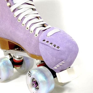 Puede incluir: Patín de ruedas de gamuza morado con cordones blancos y ruedas iridiscentes. El patín tiene un tope de puntera y una placa blanca.