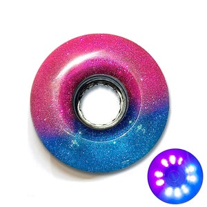 May include: A roller skate wheel with a gradient of pink to blue glitter. The wheel has a silver inner ring and a smaller, illuminated wheel with purple and blue lights.