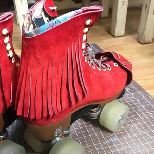 May include: A pair of red suede roller skates with fringe detailing. The skates have brown laces, white buttons, and clear wheels. The interior lining features a colorful patterned fabric. The skates are designed for recreational use.