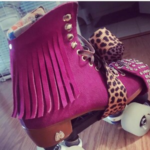 May include: Magenta roller skates with fringe detailing and gold eyelets. The skates have white wheels, brown accents, and a leopard print strap. These skates are designed for recreational use.