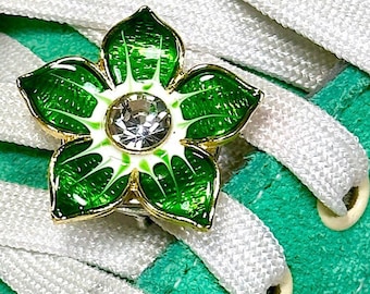 May include: Close-up of a green suede sneaker with white laces and a flower-shaped shoe charm. The charm has green enamel petals, a gold-colored border, and a clear crystal center. The laces are woven through the eyelets.