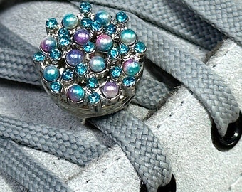May include: A close-up of a silver shoe charm with blue and iridescent beads. The charm is attached to a gray shoelace on a white shoe.