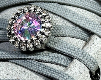 May include: Close-up of a gray sneaker with gray laces and a decorative shoe charm. The charm features a large, iridescent crystal surrounded by a silver-colored, rhinestone-studded frame, adding a touch of sparkle to the footwear.