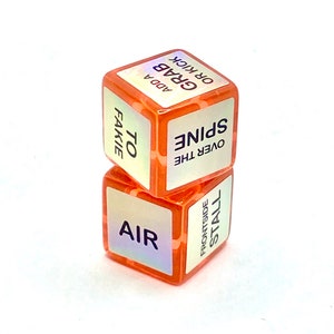 Puede incluir: Dos dados de color naranja con texto blanco. El dado superior dice "Over the Spine" y "To Fakie". El dado inferior dice "Frontside Stall" y "Air".