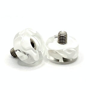 Peut inclure: Deux boutons à visser décoratifs transparents avec un motif blanc givré et tourbillonnant. Les boutons ont une forme cylindrique avec une base à vis en métal. Le fond est blanc uni.