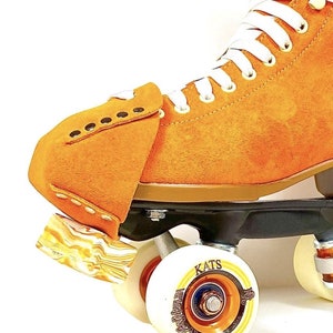 Puede incluir: Patines de ruedas de gamuza naranja con cordones blancos y ruedas negras. Las ruedas tienen un diseño dorado y negro con la palabra "KATS" impresa en ellas.