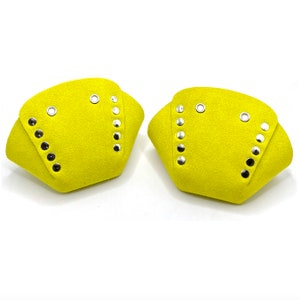 May include: Two bright yellow, suede-like fabric pieces. Each piece has a curved shape with a flap and is adorned with silver and black metal rivets and eyelets. The items are set against a white background.