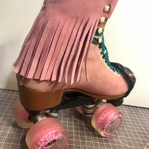 May include: Pink suede roller skate with a fringe detail. The skate has pink wheels, teal laces, and a brown heel. The top of the skate features a patterned fabric detail. A stylish roller skate.