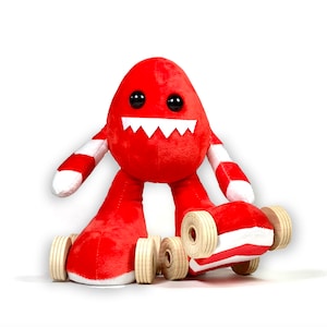 May include: A red plush monster toy with a toothy grin, wearing a pair of white and red striped socks and riding on a skateboard with four wheels.