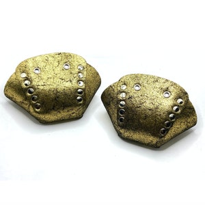 Op de afbeelding: Twee goudkleurige leren schoenbeschermers met zilveren studs. De beschermers zijn halfrond en hebben een getextureerd oppervlak.