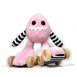 Puede incluir: Un monstruo de peluche rosa con ojos negros y una sonrisa de dientes, lleva mangas a rayas blancas y negras y patines con ruedas de madera.
