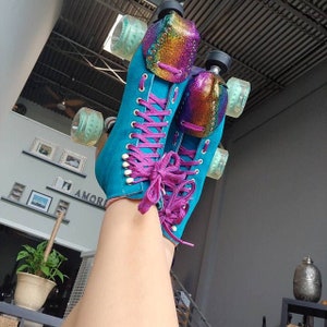 May include: A pair of teal roller skates with pink laces and iridescent rainbow toe stops. The skates are worn by a person with their legs raised in the air.