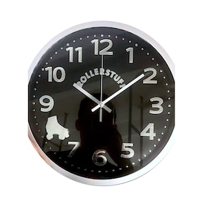 Puede incluir: Un reloj de pared negro y plateado con números y manecillas plateados. El reloj tiene un gráfico de patines y el texto "Rollerstuff" en la esfera.