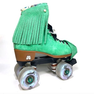 May include: A single green suede roller skate with a brown heel and black wheels. The skate has a fringe detail on the ankle and iridescent wheels. The wheels are 6.2 cm in diameter.