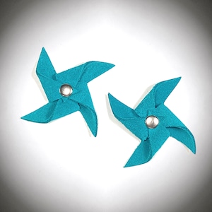 May include: Two teal blue felt pinwheels with silver metal centers. The pinwheels are on a white background.