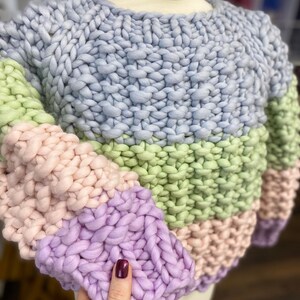 Close-up of the chunky merino wool sweater on a dress form, showing moss stitch texture, pastel stripes and the purple ribbed sleeve cuff.