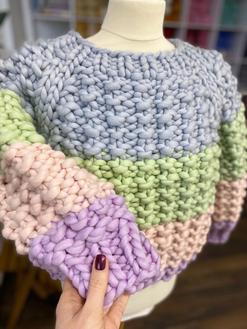 Close-up of the chunky merino wool sweater on a dress form, showing moss stitch texture, pastel stripes and the purple ribbed sleeve cuff.